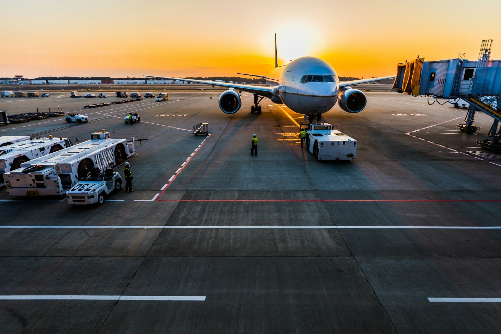 A passenger jet parked at an airport gate at sunset, with ground vehicles nearby